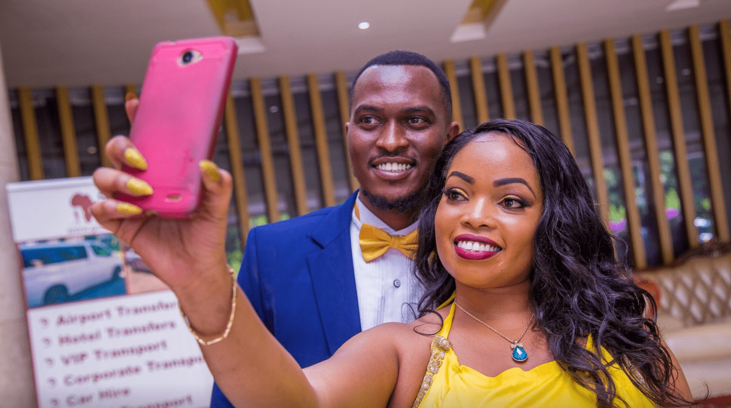 A couple in formal wear smiling and taking a selfie with a pink phone